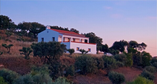 Situated on a hillside, this two-story building offers a balcony on the upper floor, surrounded by lush trees and shrubs.