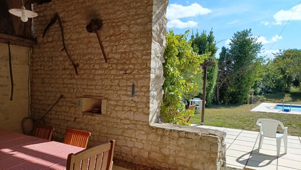 Een overdekt terras met een stenen muur, een tafel met geruit tafelkleed, houten stoelen en uitzicht op een zwembad en groen.