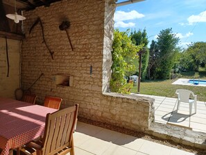 A covered patio with a stone wall, a table with a checkered tablecloth, wooden chairs, and a view of a swimming pool and greenery. - Spacious Farmhouse with Private Pool. Ideal for 10 adults. (Jaulnay)