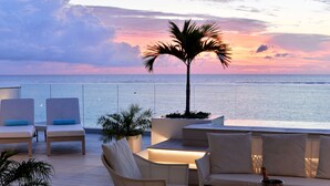 Una terraza con asientos blancos acolchados, una palmera, una mesa con una botella, vasos y flores.