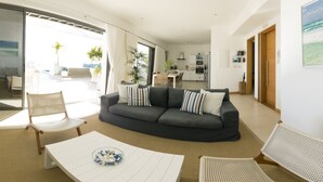 Una sala de estar moderna con un sofá gris oscuro, cojines blancos y azules estampados, una mesa de centro blanca, sillas de mimbre y vistas a la cocina y a un área exterior a través de una puerta de vidrio.