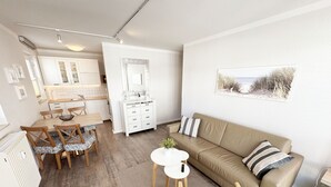 Moderni olohuone, jossa on beigenvärinen sohva, ruokapöytä tuoleineen, pieni valkoinen pöytä, jossa on kasvi, ja taustalla keittiö.