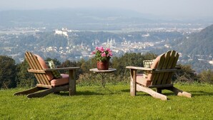 Zwei Adirondack-Stühle mit karierten Kissen und ein kleiner Tisch mit einem Blumentopf, mit Blick auf eine malerische Aussicht auf eine Stadt und eine Burg.