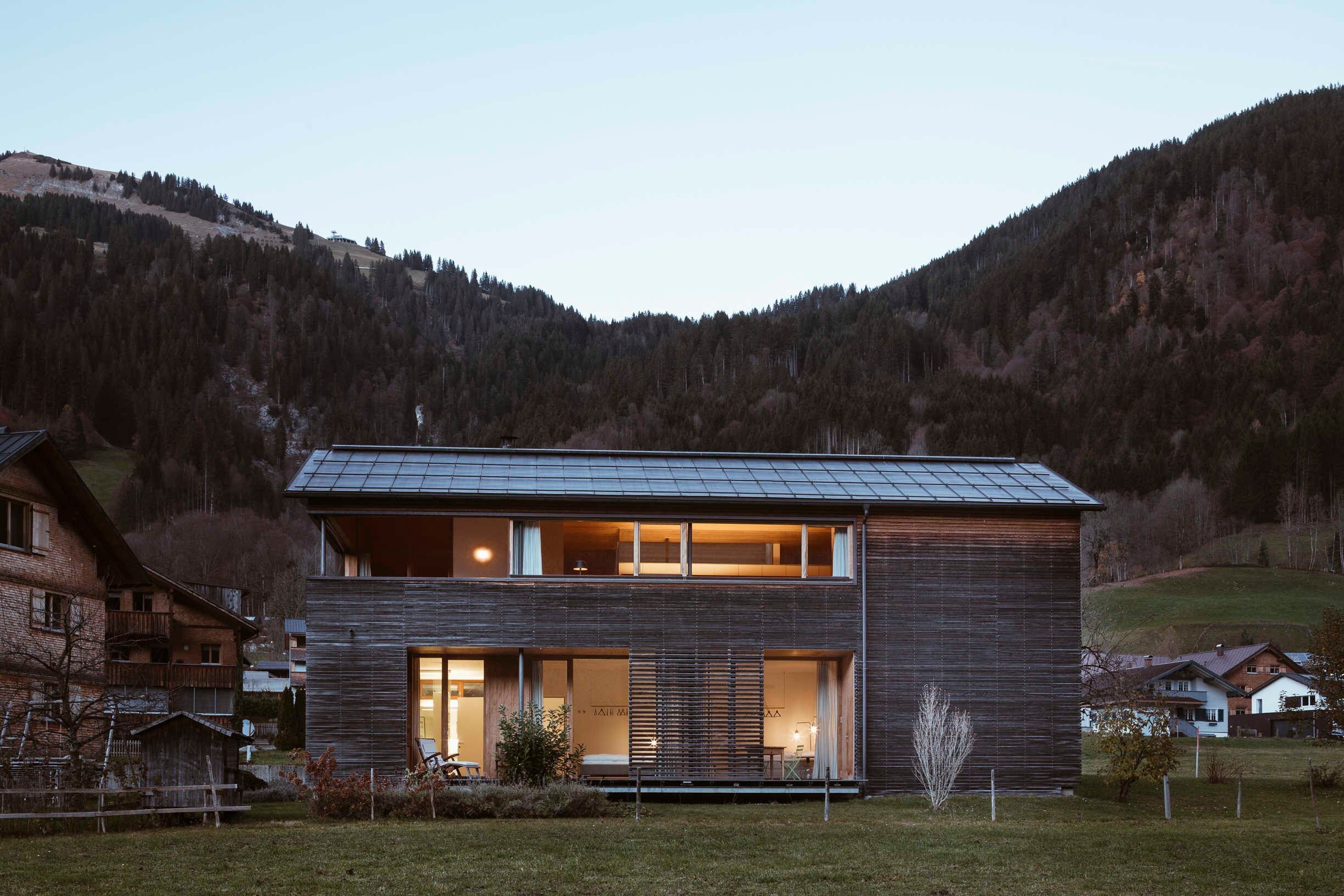 Een modern huis met twee verdiepingen, grote ramen en een schuin dak, omgeven door een bergachtig landschap.