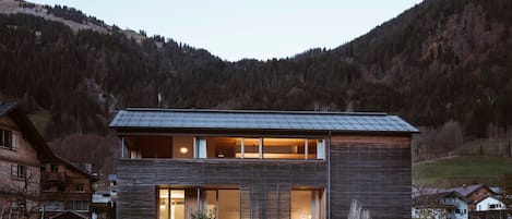 Una casa moderna de dos pisos con ventanas amplias y techo inclinado, rodeada por un paisaje montañoso.