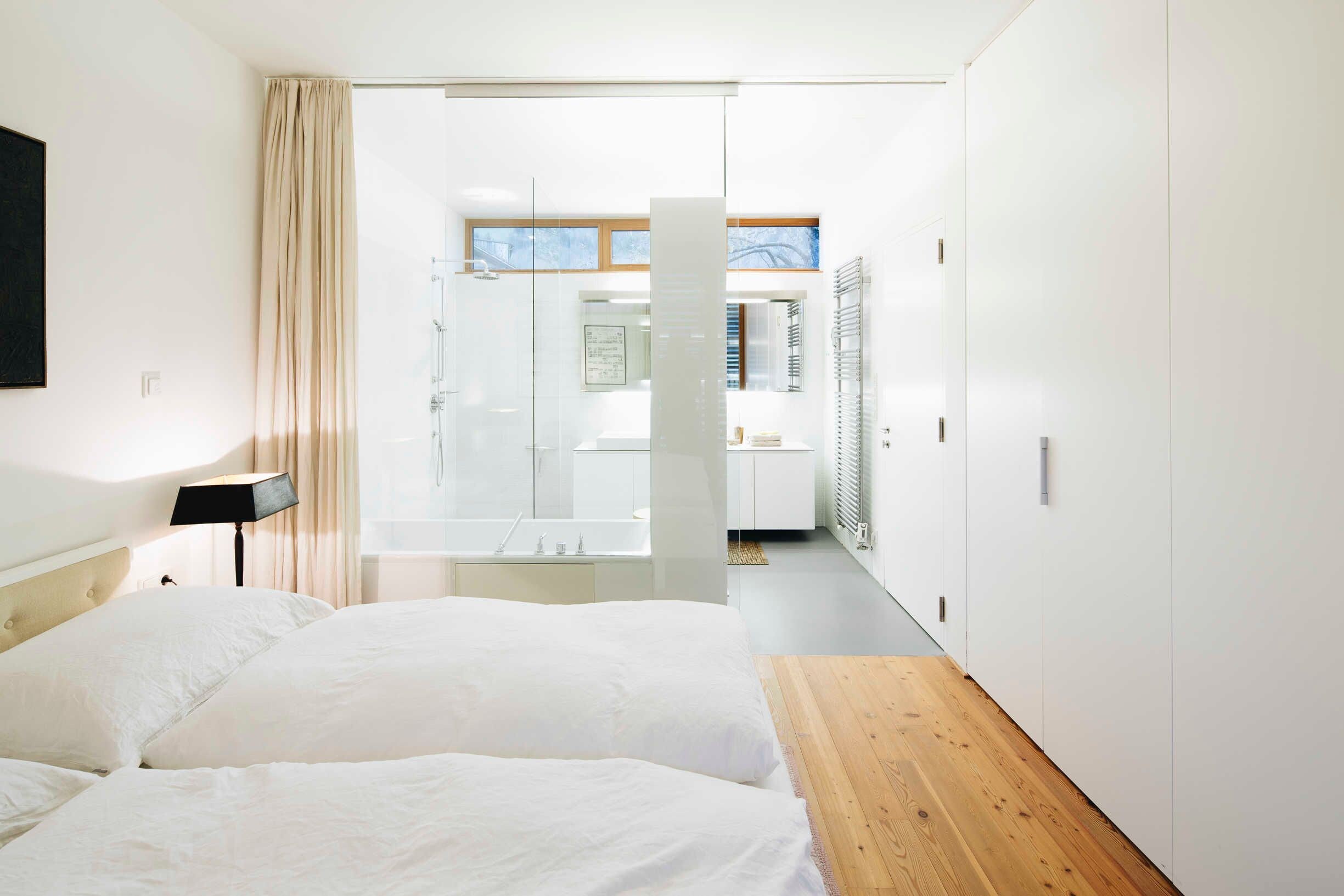 A modern hotel room with a large bed, a wooden bench, a glass door leading to a bathroom, and a view of the outside.