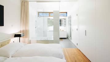 A modern hotel room with a large bed, a wooden bench, a glass door leading to a bathroom, and a view of the outside.