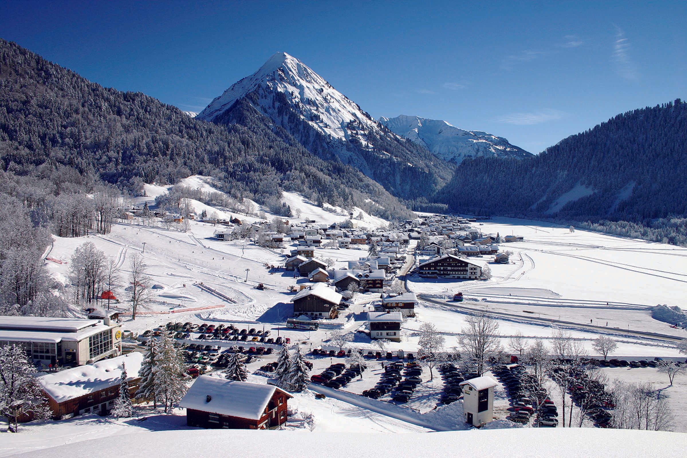 A snow-covered mountain town with buildings, a ski slope, and a parking lot.