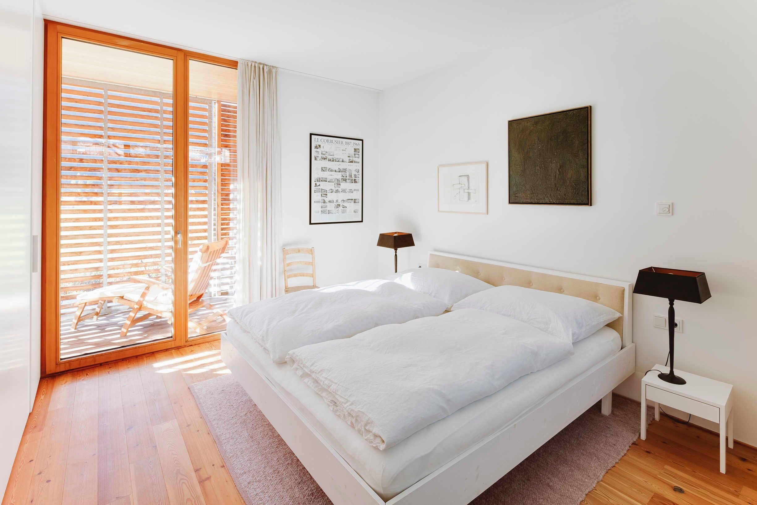 Une chambre à coucher avec un lit, une table de chevet, une lampe, un tableau encadré et une fenêtre avec des stores.