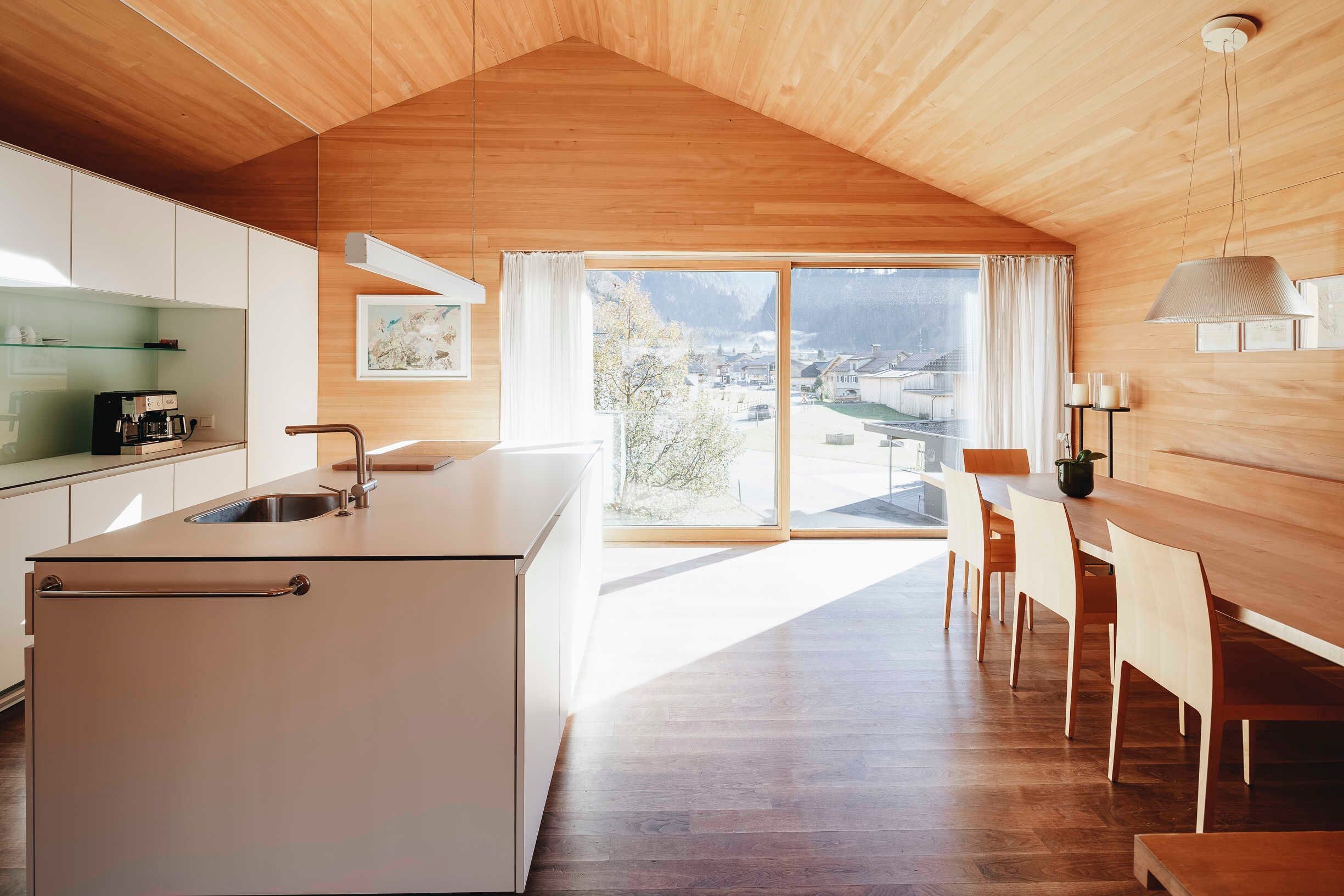 A modern kitchen with a large island, a dining area with wooden chairs, and a view of a town through large windows.
