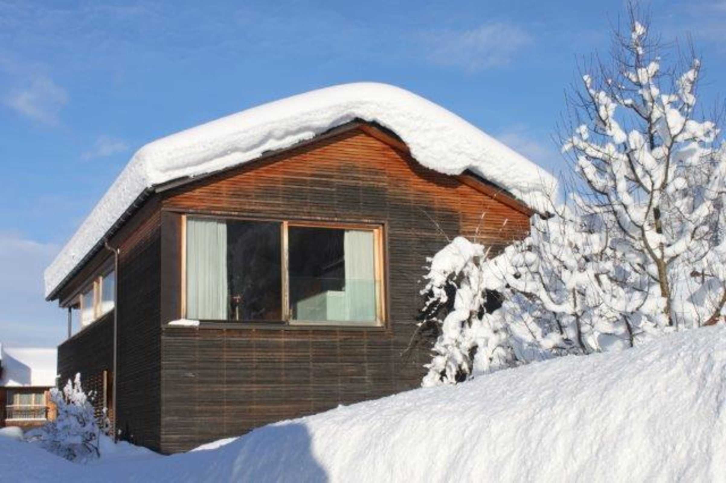 A wooden cabin with a snow-covered roof, a large window, and a snow-covered tree in the foreground.