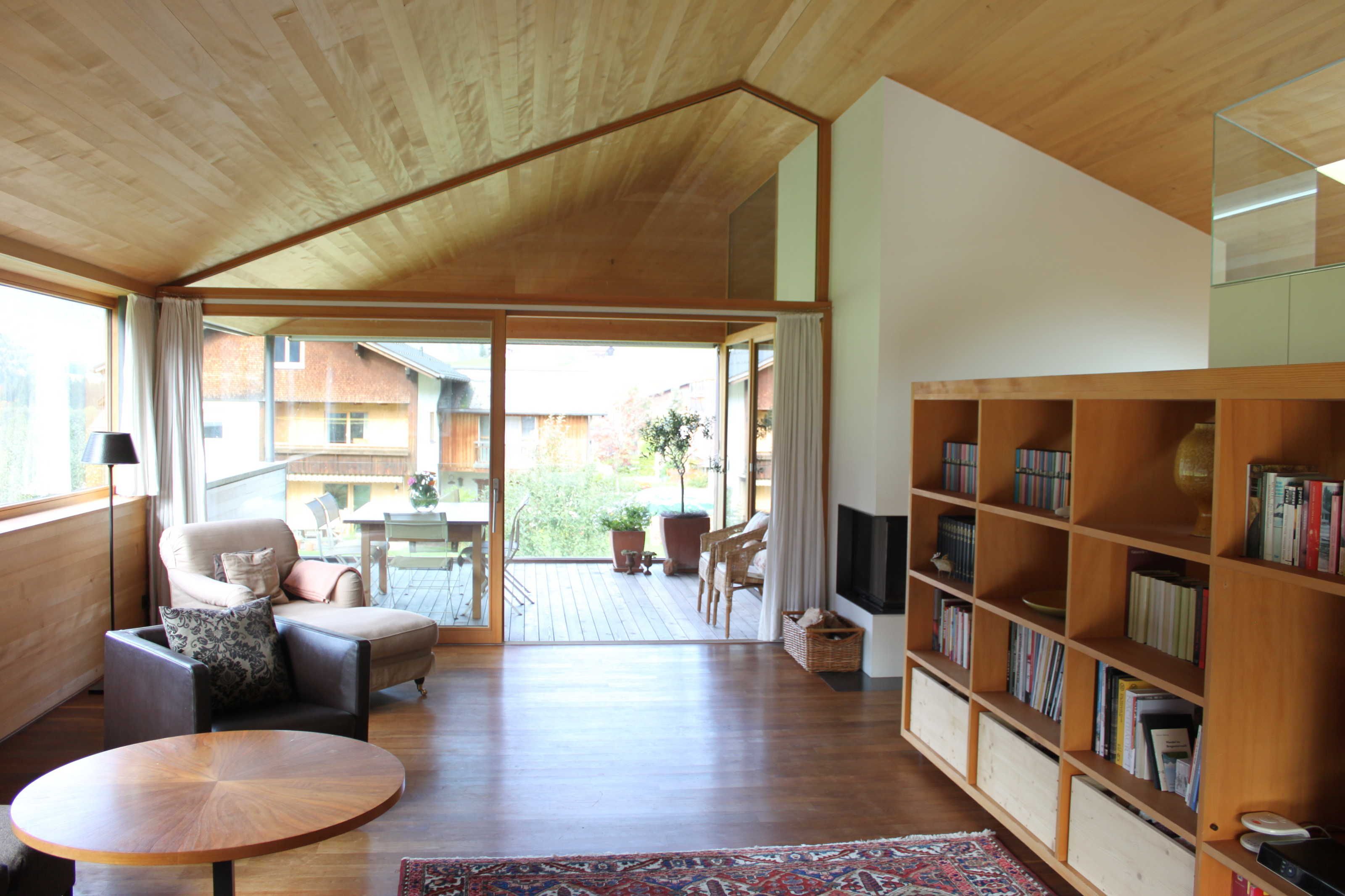 A modern living room with a wooden ceiling, a sofa, a round table, a bookshelf, and a view through a glass door to an outdoor patio.
