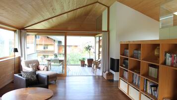 A modern living room with a wooden ceiling, a sofa, a round table, a bookshelf, and a view through a glass door to an outdoor patio.