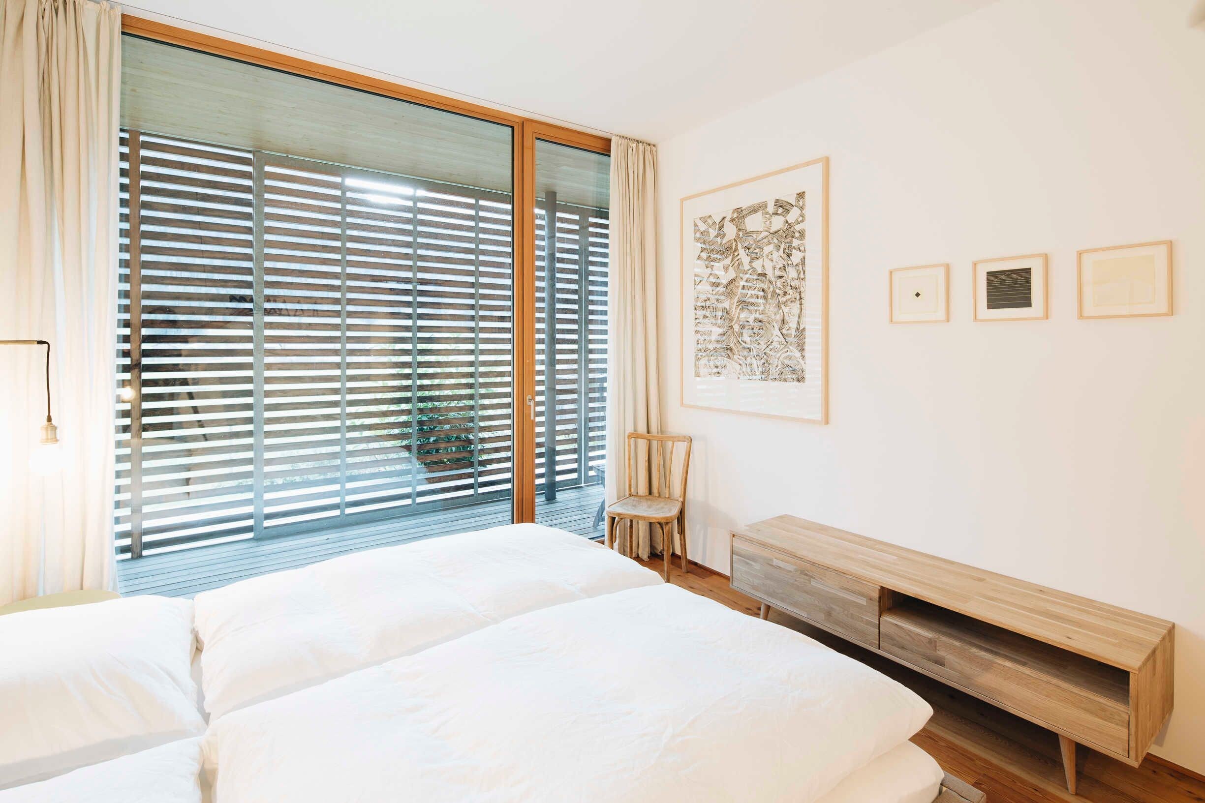 Een slaapkamer met een grote schuifdeur, een bed, een stoel, een houten nachtkastje en een lijst met een kunstwerk aan de muur.