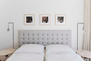 Een slaapkamer met een gestoffeerd hoofdeinde, twee bedden, nachtkastjes en ingelijste foto's aan de muur.