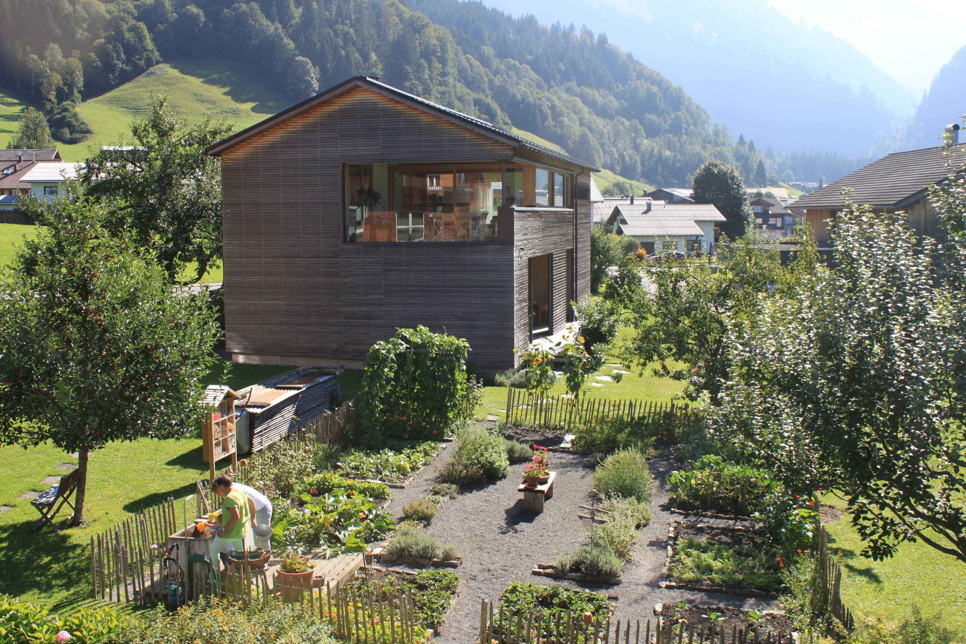Een modern huis met een groot raam, omringd door een verzorgde tuin en een berg op de achtergrond.