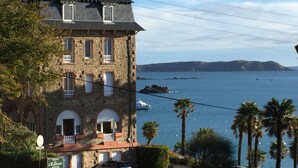 Ein Gebäude aus Stein mit Bogenfenstern und einem Balkon, mit Blick auf eine Küstenlandschaft mit Palmen und freiem Blick auf das Meer.