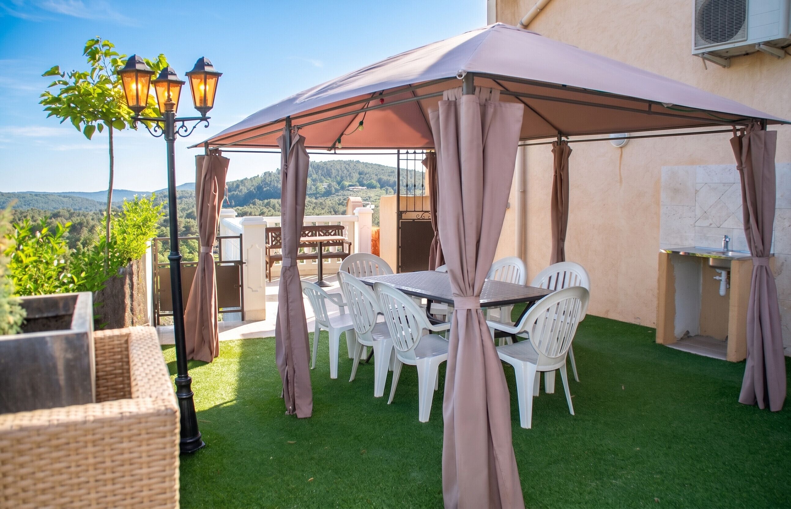 Um pátio externo com mesa e cadeiras, uma área coberta com cortinas, e vista para as colinas.