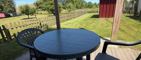 A wooden deck with a round table and chairs, a wooden fence, and a red shed in a grassy area.