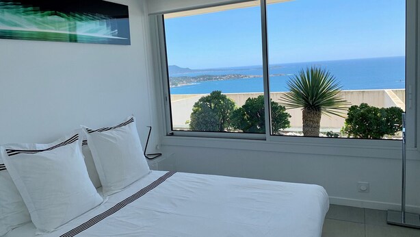 A bedroom with a large window offering a view of the sea and distant land.