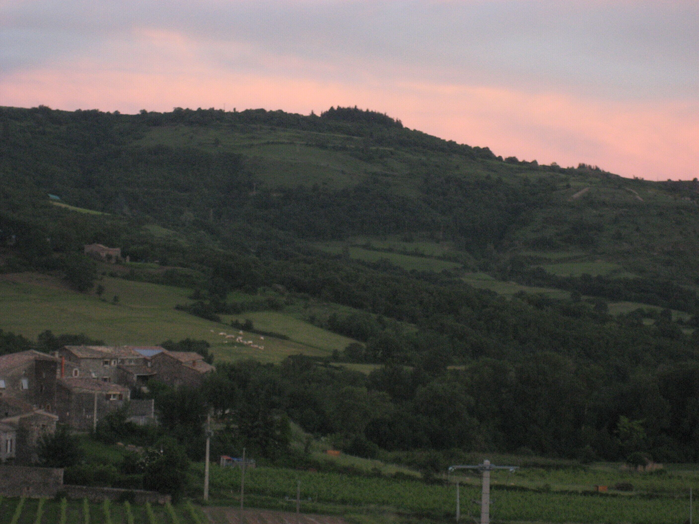 Un paysage rural caractérisé par des collines douces, un petit groupe de bâtiments et quelques arbres dispersés.