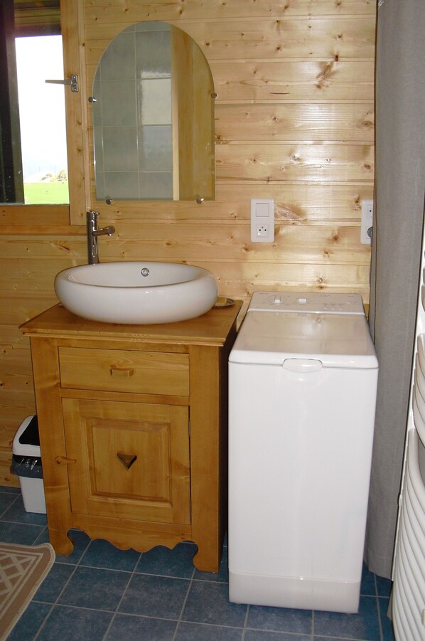 Une salle de bain rustique, aménagée dans une cabane en bois, avec un lavabo blanc, un miroir et un lave-linge.
