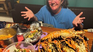 A person seated at a table with a large lobster, a bowl of salad, and a pot of food. - Your Tropical Beach Vacation! (Morgans Bluff)