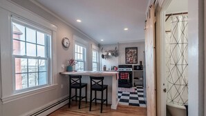 A kitchen with a wooden floor, two bar stools, a window, a clock, and a microwave. - Vintage Maine & Modern Convenience in Portlands Exclusive East End (Portland)
