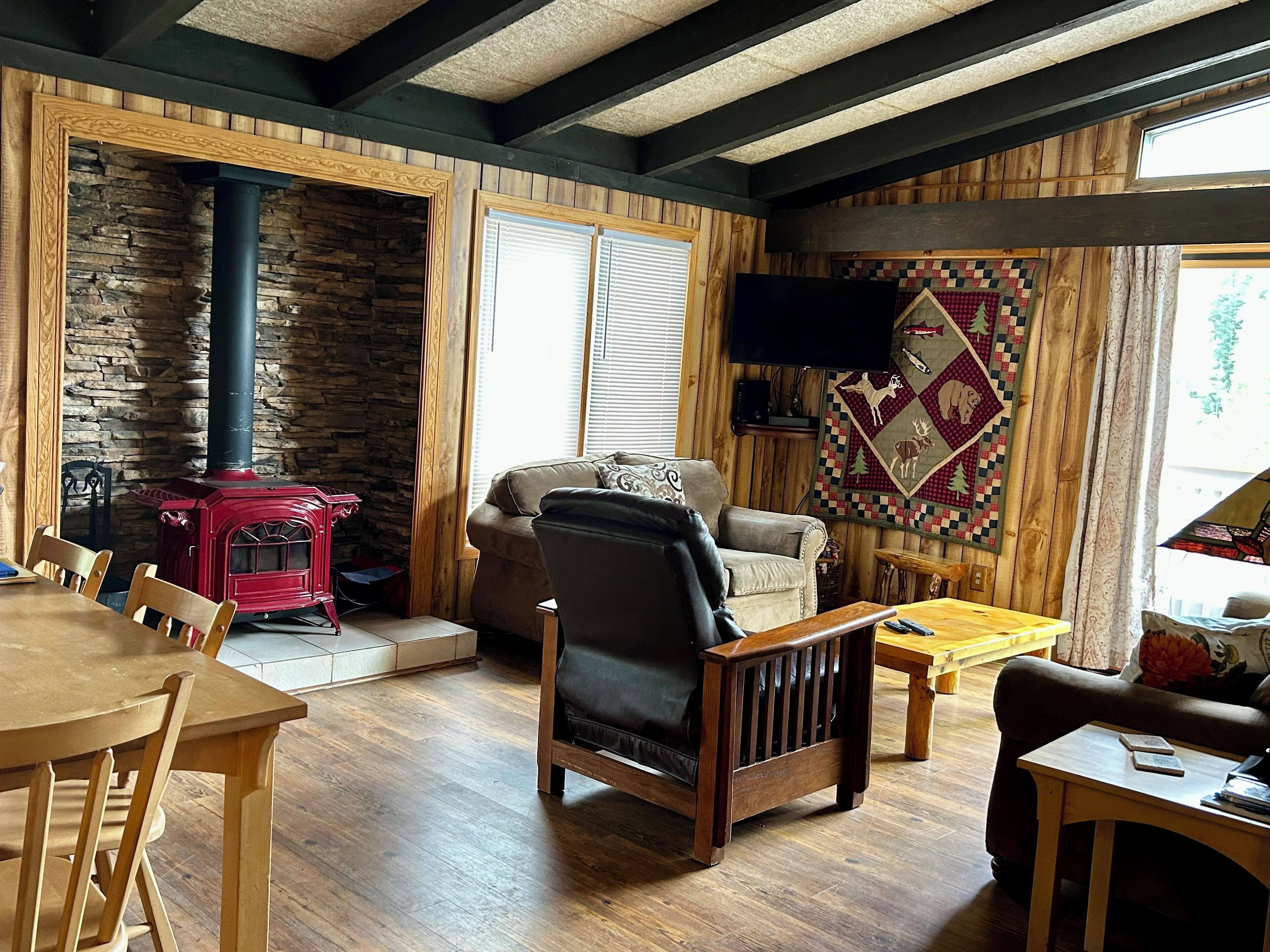 Un salón acogedor con pared de piedra, vigas de madera, estufa roja, sofá de cuero y un tapiz acolchado.