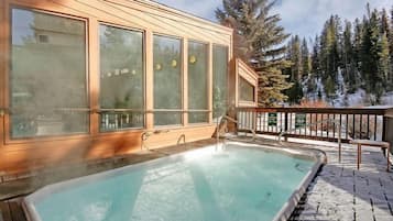Una cabaña de madera con un jacuzzi en una terraza de madera, rodeada de árboles cubiertos de nieve.