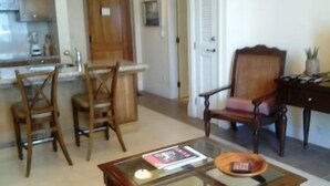 Un salon avec une table basse en verre, des chaises en bois et un coin repas avec une cuisine en arrière-plan.