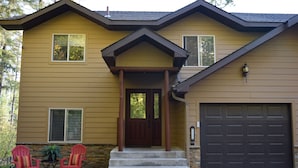Une maison à deux étages avec un porche à l’avant, un garage et un petit patio agrémenté de deux chaises Adirondack rouges et d’une plante en pot.