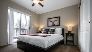 Une chambre avec un grand lit, deux tables de chevet, un ventilateur de plafond et une fenêtre donnant sur des arbres.