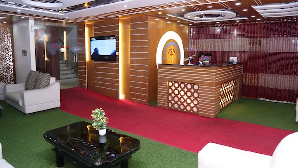 A hotel reception area with a red carpet, a reception desk, a sofa, and a television screen. - Platinum Hotel Ltd (Dhaka)