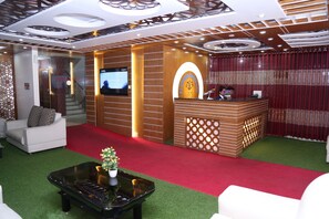 A hotel reception area with a red carpet, a reception desk, a sofa, and a television screen. - Platinum Hotel Ltd (Dhaka)
