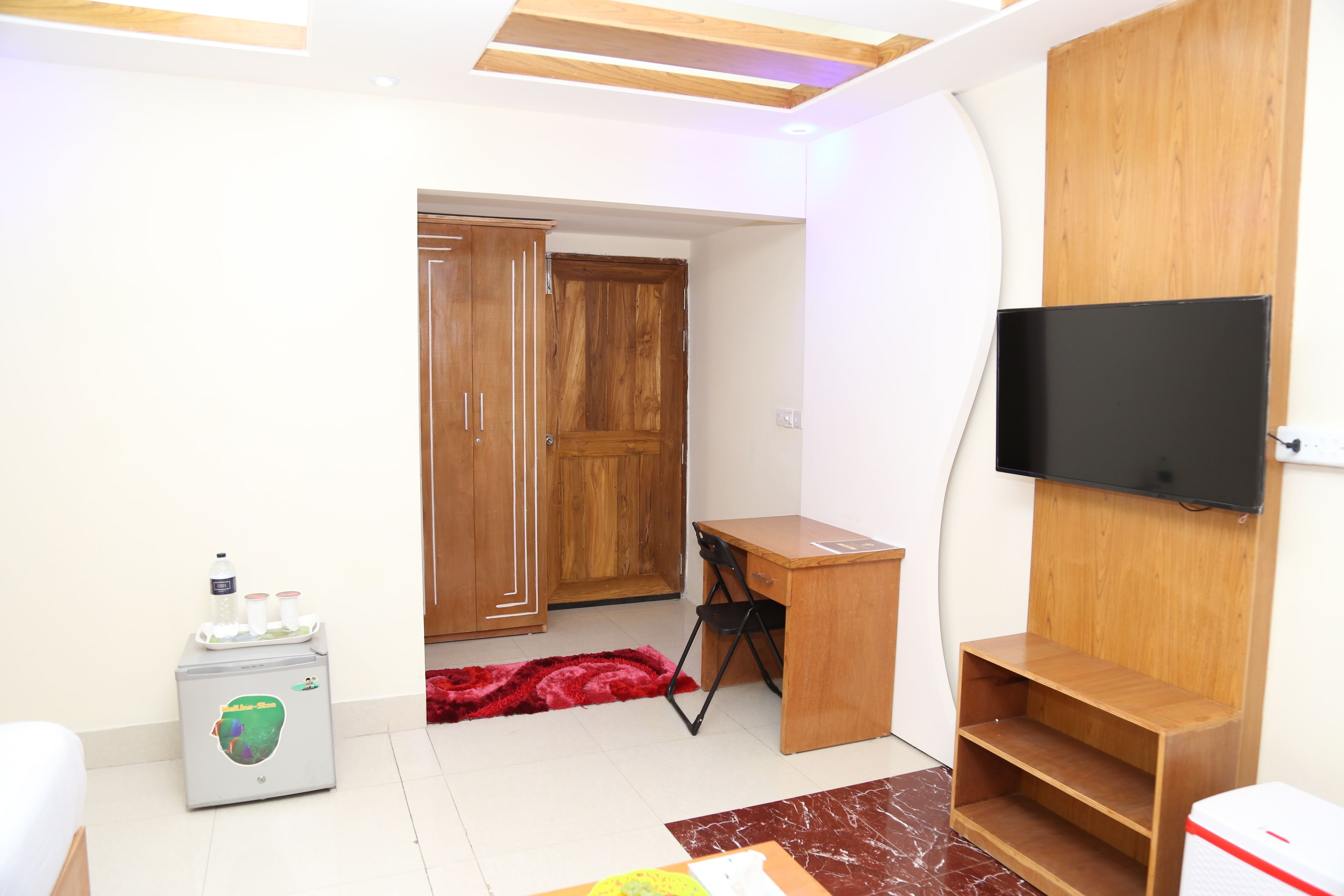 A room with a wooden desk, a television mounted on the wall, a wardrobe, and a small refrigerator.