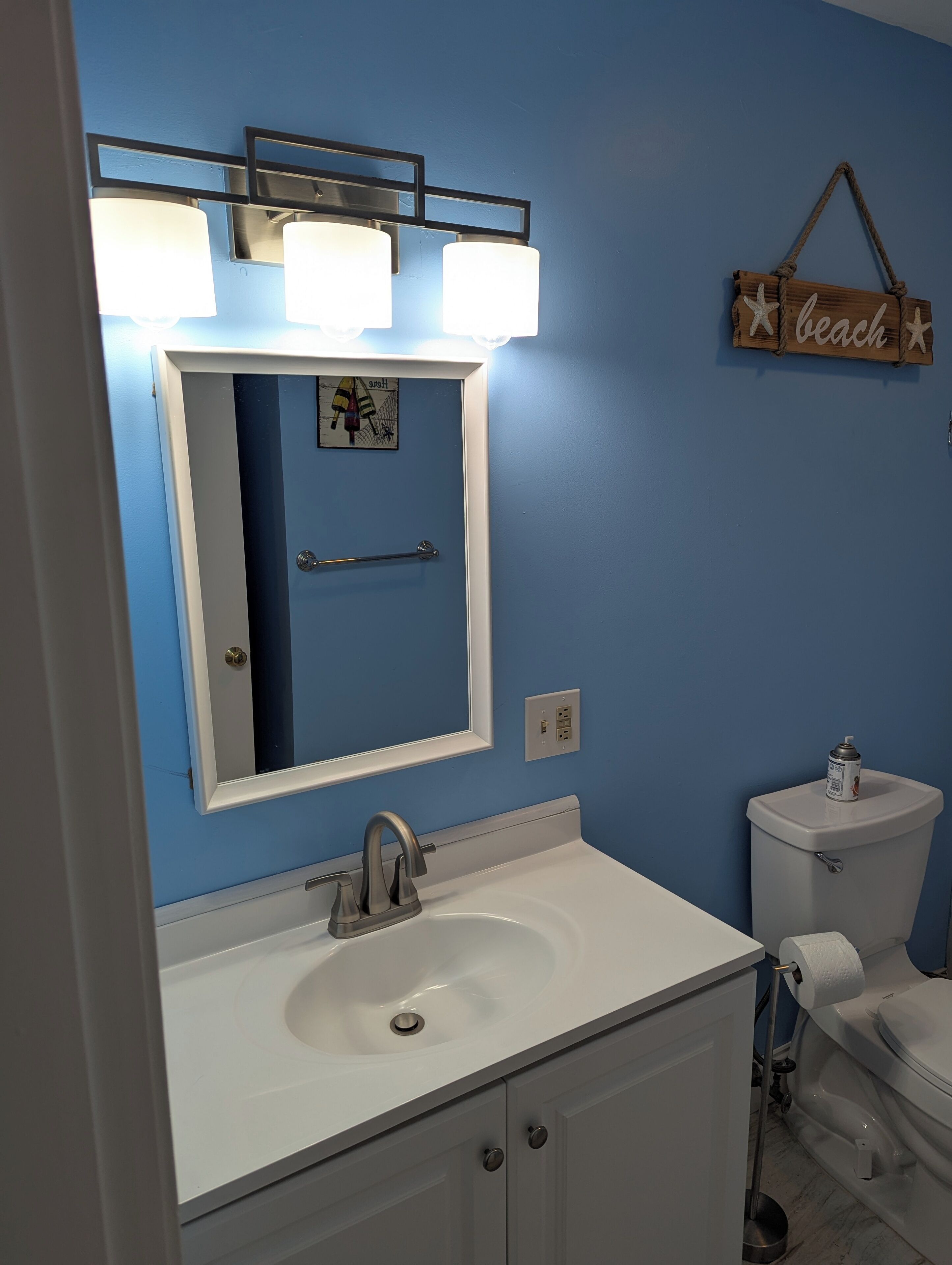 A bathroom with a white sink, a mirror, and a toilet. There is a wall-mounted light fixture above the mirror and a small wooden sign on the wall.