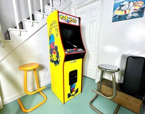 Uma máquina de jogos eletrônicos amarela com a inscrição 'PAC-MAN', ao lado de uma escada e dois banquinhos.