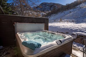 A hot tub with a wooden exterior, set against a snowy landscape with mountains in the background.