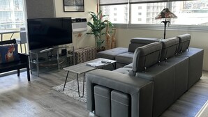 Ein modernes Wohnzimmer mit einer Eckcouch, einem Flachbildfernseher, einem Deckenventilator und großen Fenstern mit Jalousien.