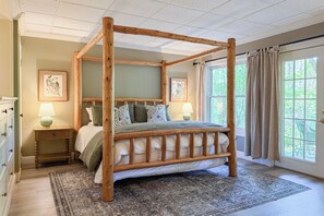 A four-poster bed with a wooden frame, two bedside tables with lamps, a patterned rug, and a view of greenery through a window.