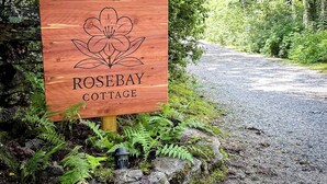 Un segnale di legno per Rosebay Cottage con un motivo floreale, situato accanto a un sentiero sterrato immerso nel verde.
