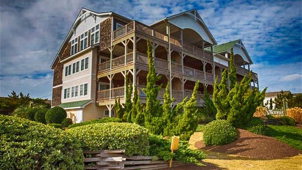 A large, multi-story building with a prominent balcony area, surrounded by well-maintained greenery and a clear sky.