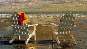 Two Adirondack chairs on a sandy beach with a view of the ocean.