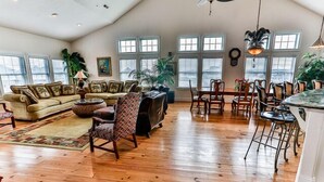 A spacious living room with a high ceiling, wooden flooring, a sofa set, a dining area with chairs, and large windows providing natural light.