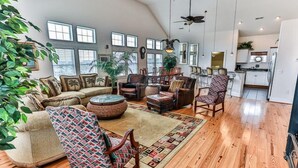 A spacious living room with a high ceiling, wooden flooring, and a large window. There is a dining area with wooden furniture and a kitchen in the background.