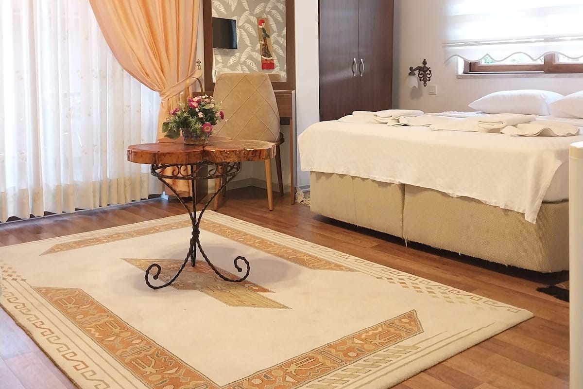 Une chambre d’hôtel avec un lit, une table en bois sur laquelle se trouve un vase, une chaise, une armoire et une grande fenêtre avec des rideaux.