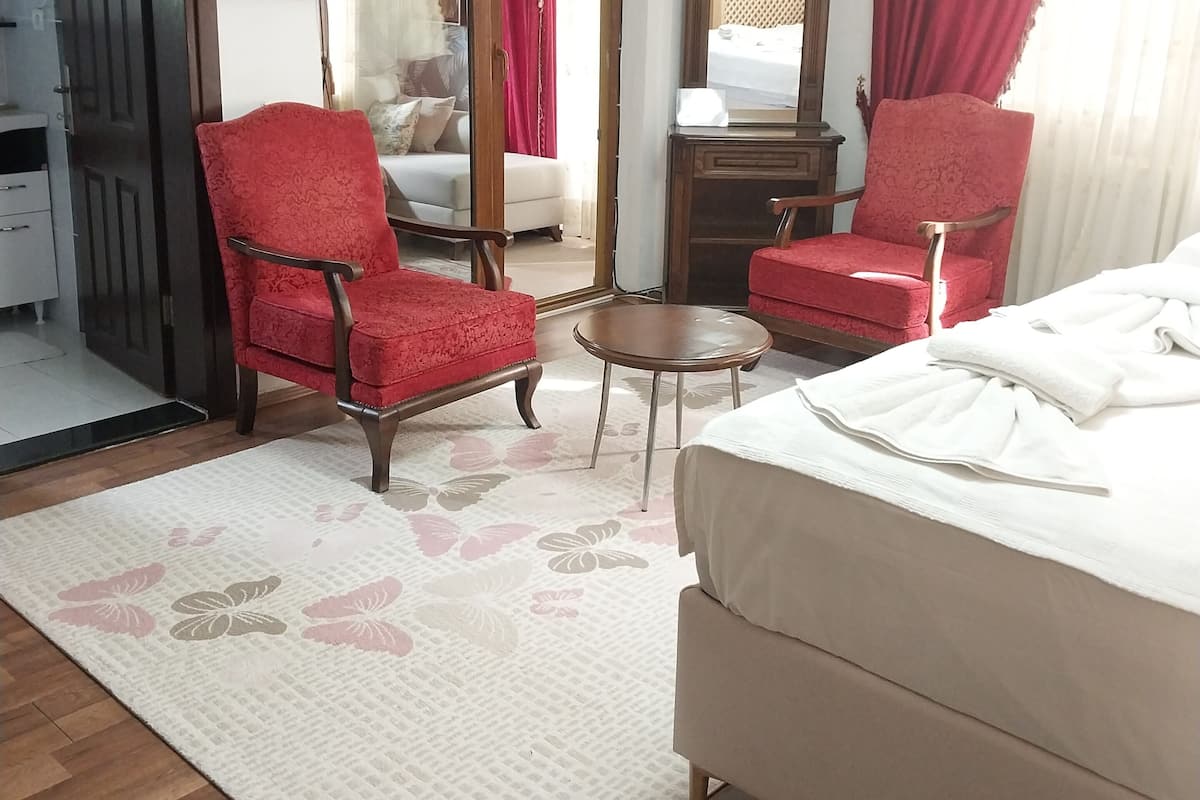 Une chambre d’hôtel avec un lit, deux fauteuils rouges, une petite table ronde, une commode et un miroir.