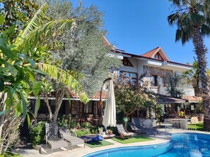 A pool area with lounge chairs, a pool, and a house with a tiled roof and stone facade. - Jasmin Apart Hotel (Fethiye)