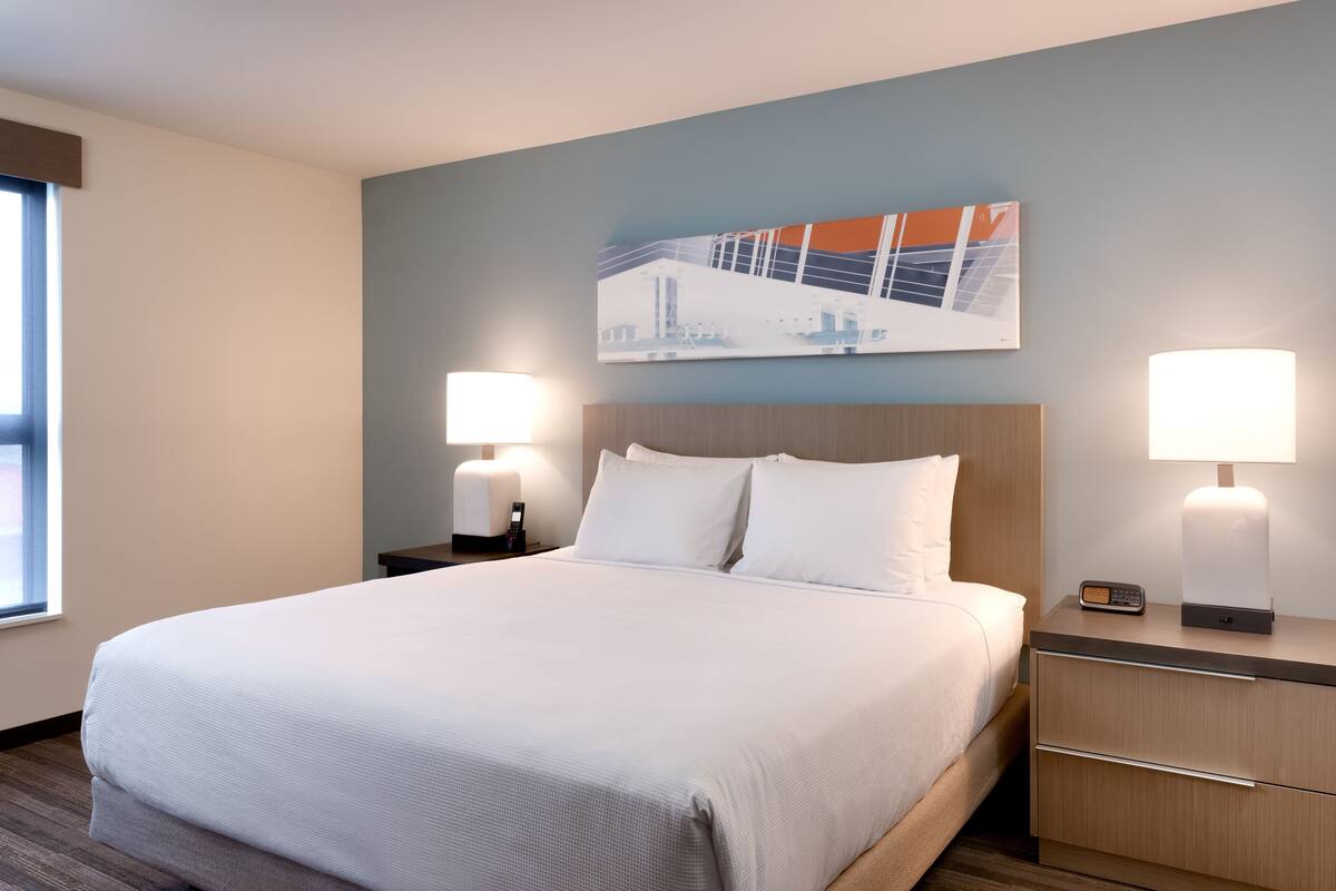 Ein Hotelzimmer mit einem großen Bett, zwei Nachttischen mit Lampen, einem Beistelltisch, einem Bild an der Wand und einem Fenster mit Jalousien.